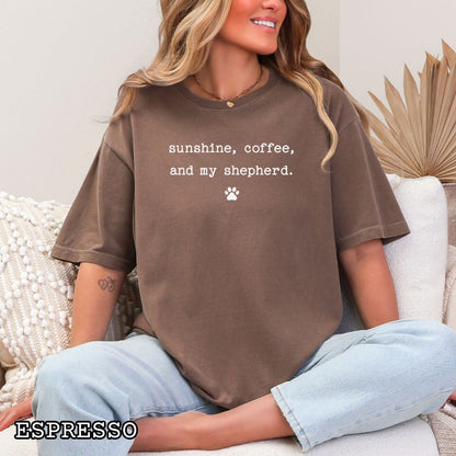 Brown Comfort Colors t-shirt for German Shepherd lovers with 'sunshine, coffee, and my shepherd' text in minimalist typewriter lettering and paw print, shown on a wooden surface with accessories.