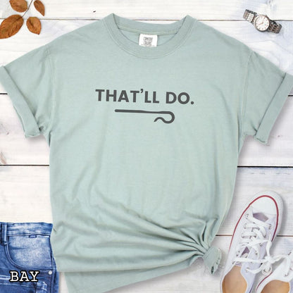 Light green Comfort Colors t-shirt for  sheep herding dog trainers with 'THAT'LL DO.' text and a shepherd's crook graphic, shown on a wooden surface with jeans and sneakers.
