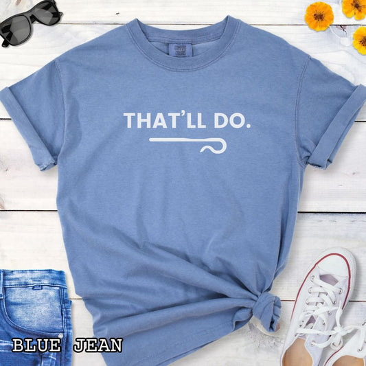 Blue Comfort Colors t-shirt for herding dog trainers with 'THAT'LL DO.' text and a shepherd's crook graphic, shown on a wooden surface with jeans and sneakers.