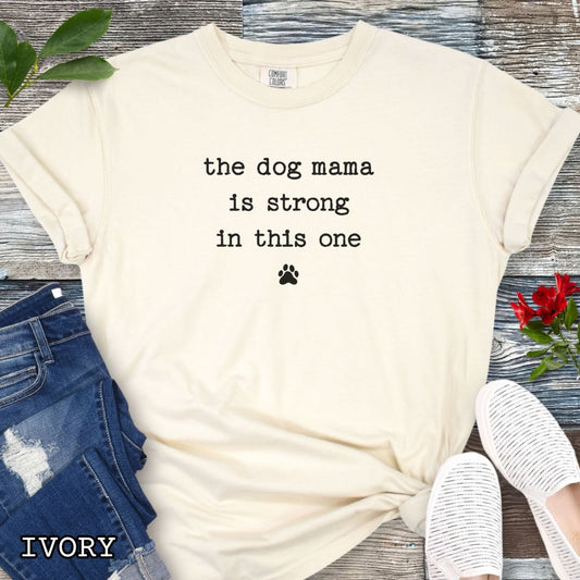 Beige Comfort Colors t-shirt for dog moms with the humorous text 'the dog mama is strong in this one' in minimalist typewriter lettering, shown on a wooden surface with denim jeans and flowers.