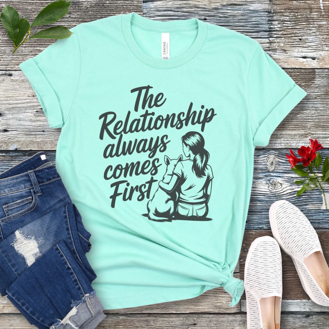 A mint green t-shirt for dog lovers and dog trainers with an illustration of a girl sitting with her dog, and the text 'The Relationship always comes First', displayed with jeans and shoes.
