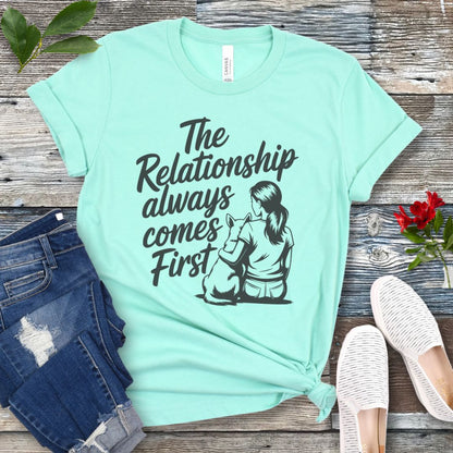 A mint green t-shirt for dog lovers and dog trainers with an illustration of a girl sitting with her dog, and the text 'The Relationship always comes First', displayed with jeans and shoes.