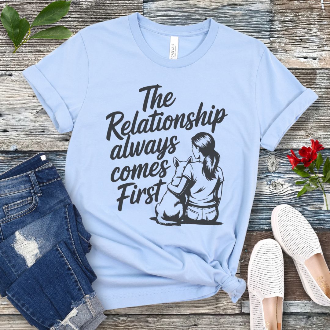 A light blue t-shirt for dog lovers and dog trainers with an illustration of a girl sitting with her dog, and the text 'The Relationship always comes First', displayed with jeans and shoes.