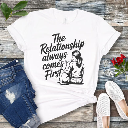 A white t-shirt for dog lovers and dog trainers with an illustration of a girl sitting with her dog, and the text 'The Relationship always comes First', displayed with jeans and shoes.