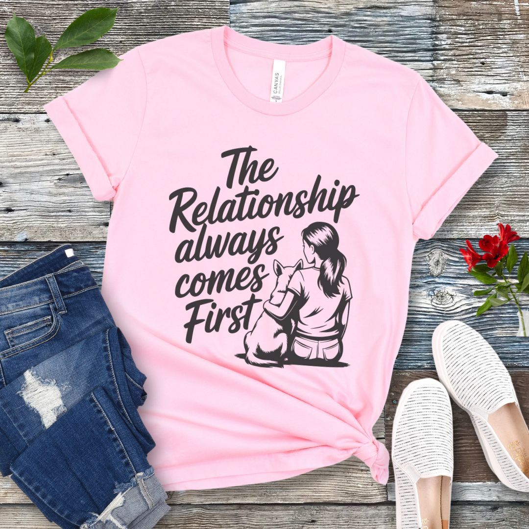 A pink t-shirt for dog lovers and dog trainers with an illustration of a girl sitting with her dog, and the text 'The Relationship always comes First', displayed with jeans and shoes.