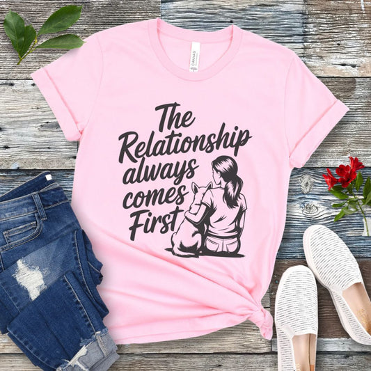 A pink t-shirt for dog lovers and dog trainers with an illustration of a girl sitting with her dog, and the text 'The Relationship always comes First', displayed with jeans and shoes.