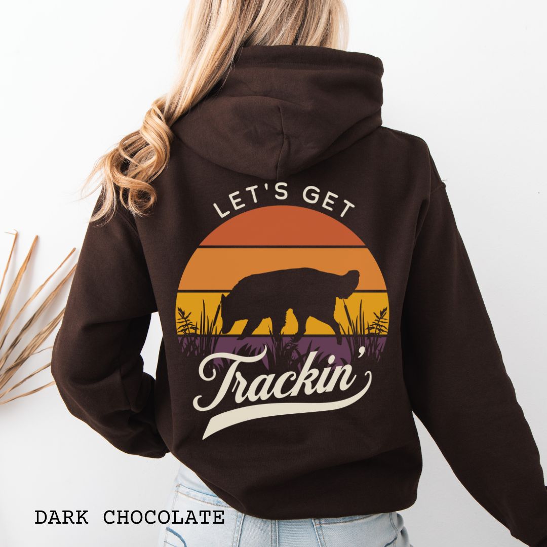 A woman shown from the back wearing a dark brown hoodie with a large retro sunset graphic on the back. The graphic features a custom breed tracking dog silhouette, with the words 'LET'S GET Trackin' in vintage retro script.
