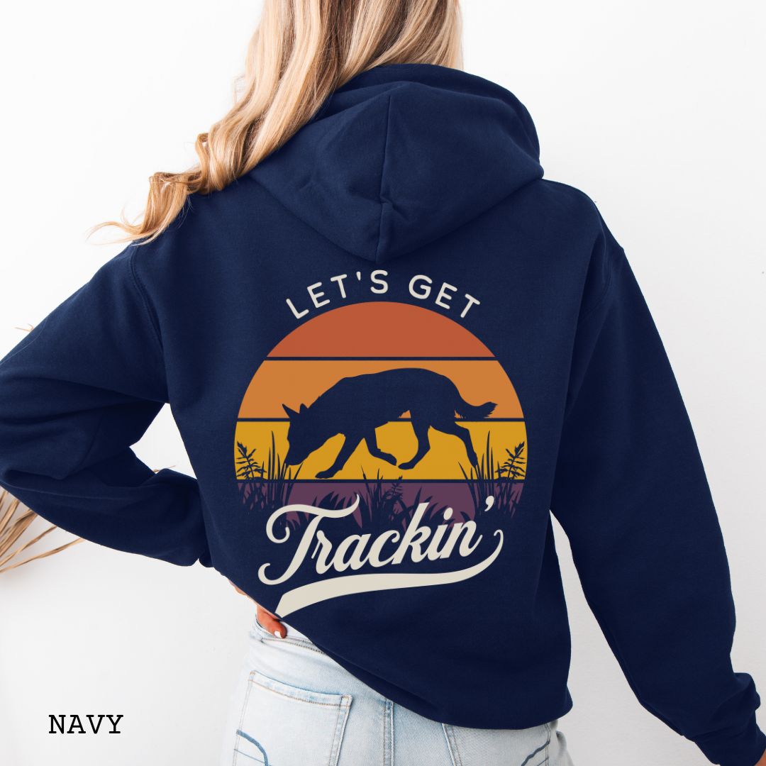 A woman shown from the back wearing a navy blue hoodie with a large retro sunset graphic on the back. The graphic features a custom breed tracking dog silhouette, with the words 'LET'S GET Trackin' in vintage retro script.