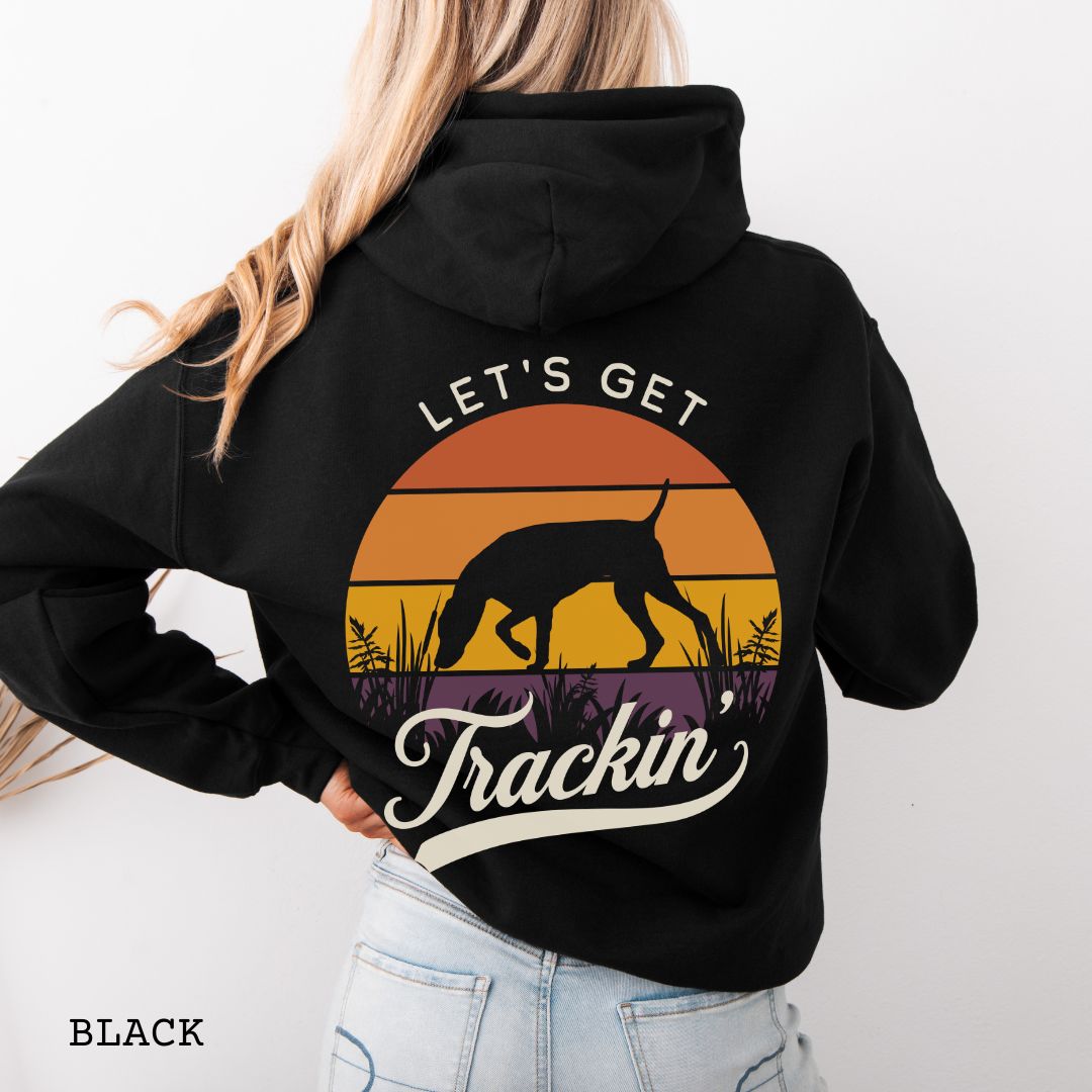 A woman shown from the back wearing a black hoodie with a large retro sunset graphic on the back. The graphic features a custom breed tracking dog silhouette, with the words 'LET'S GET Trackin' in vintage retro script.