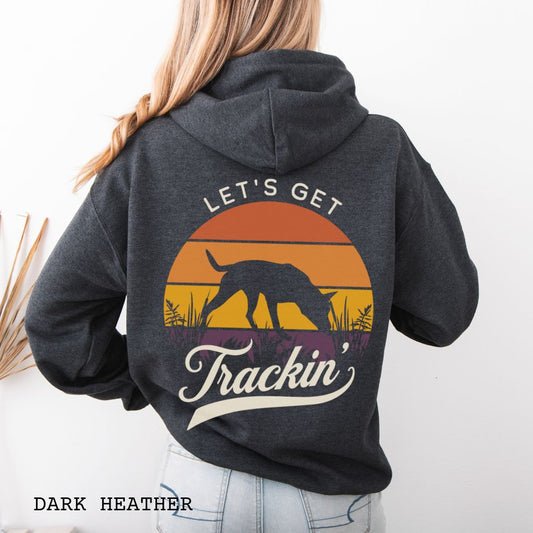A woman shown from the back wearing a dark heather gray hoodie with a large retro sunset graphic on the back. The graphic features a custom breed tracking dog silhouette, with the words 'LET'S GET Trackin' in vintage retro script.