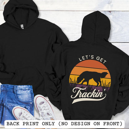 Two black hoodies shown in a flat-lay arrangement on a white wood background, with jeans and white sneakers as styling props. The front hoodie is plain black, and the back hoodie features a large retro sunset-style graphic with a dog silhouette and the text 'LET'S GET Trackin''.