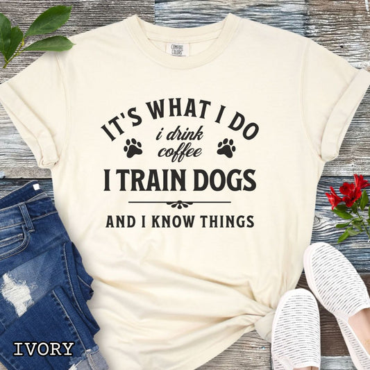 Beige (Ivory) Comfort Colors dog training t-shirt with text 'It's what I do, I drink coffee, I train dogs, and I know things' on a wooden surface with denim shorts and a flower.