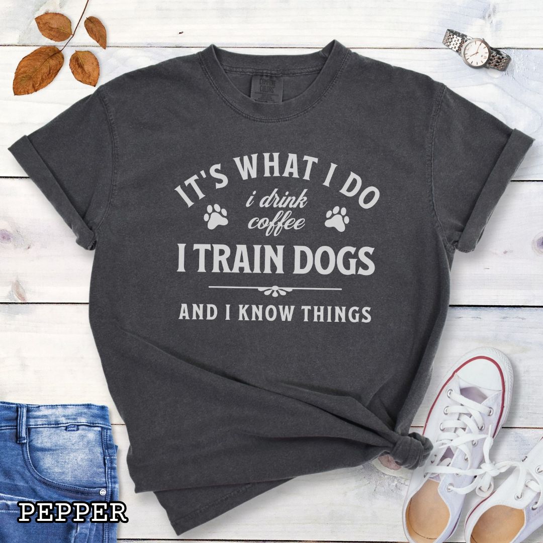 Dark grey (Pepper) Comfort Colors dog training t-shirt with text 'It's what I do, I drink coffee, I train dogs, and I know things' on a wooden surface with denim shorts and a flower.