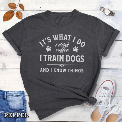 Dark grey (Pepper) Comfort Colors dog training t-shirt with text 'It's what I do, I drink coffee, I train dogs, and I know things' on a wooden surface with denim shorts and a flower.