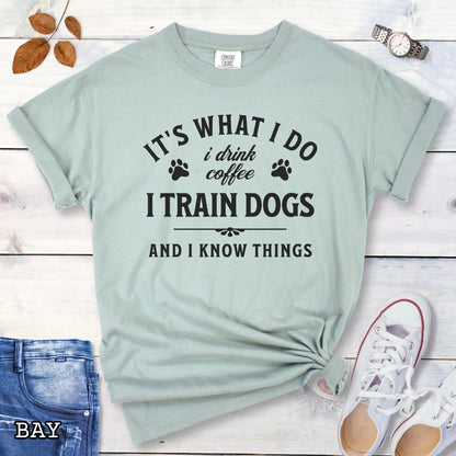 Light green (Bay) Comfort Colors dog training t-shirt with text 'It's what I do, I drink coffee, I train dogs, and I know things' on a wooden surface with denim shorts and a flower.