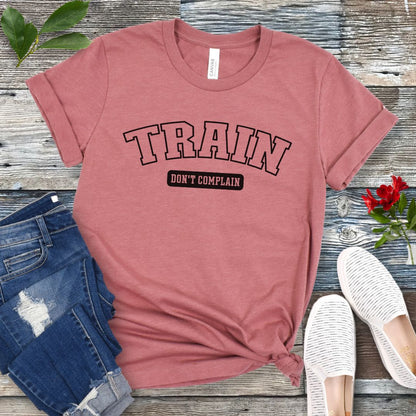 A mauve dog training t-shirt with the text 'TRAIN DON'T COMPLAIN' in athletic style lettering, presented flat with a pair of blue jeans and white shoes.