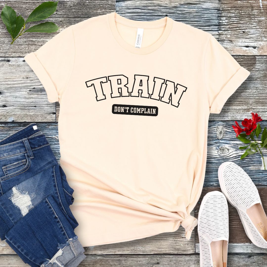A beige dog training t-shirt with the text 'TRAIN DON'T COMPLAIN' in athletic style lettering, presented flat with a pair of blue jeans and white shoes.