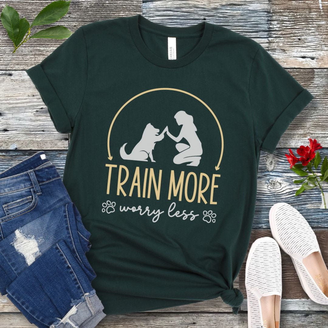 A dark green dog training t-shirt with a graphic of a dog and a person silhouetted in a circular frame, and text that reads 'TRAIN MORE WORRY LESS'. The t-shirt is presented on a wooden background with a pair of blue jeans and white shoes.
