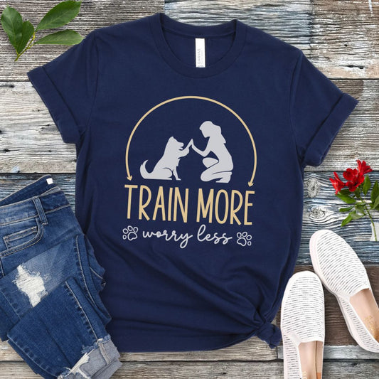 A navy blue dog training t-shirt with a graphic of a dog and a person silhouetted in a circular frame, and text that reads 'TRAIN MORE WORRY LESS'. The t-shirt is presented on a wooden background with a pair of blue jeans and white shoes.