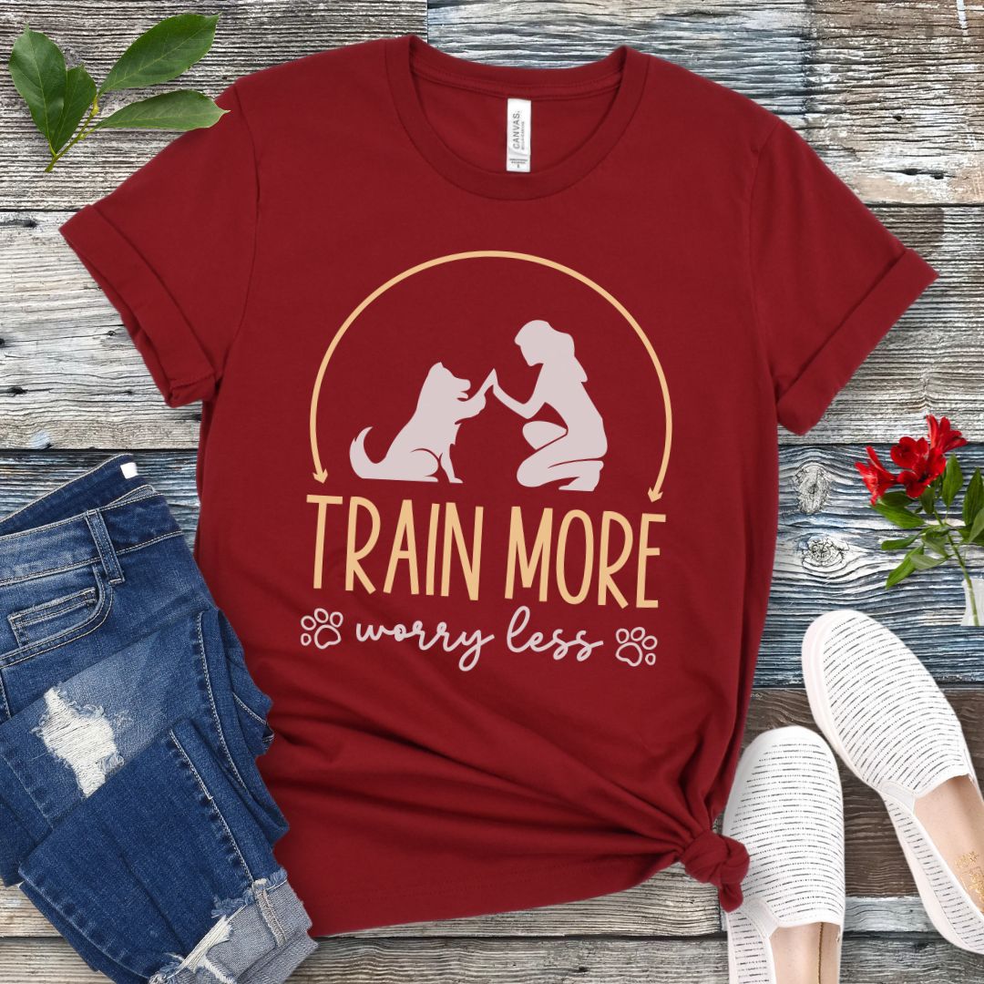 A dark red dog training t-shirt with a graphic of a dog and a person silhouetted in a circular frame, and text that reads 'TRAIN MORE WORRY LESS'. The t-shirt is presented on a wooden background with a pair of blue jeans and white shoes.
