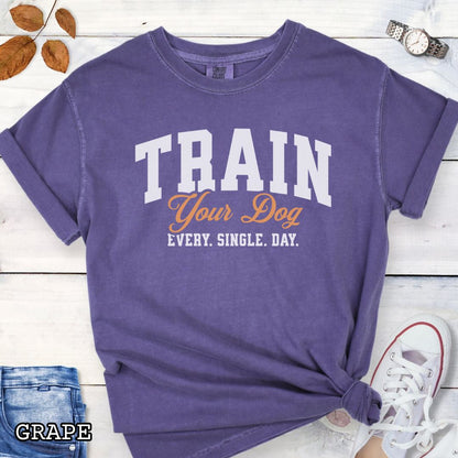 Purple Comfort Colors t-shirt for dog training enthusiasts with 'TRAIN Your Dog EVERY. SINGLE. DAY.' text in bold athletic style lettering, shown on a wooden surface with jeans and shoes.