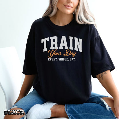 Black Comfort Colors t-shirt for dog training enthusiasts with 'TRAIN Your Dog EVERY. SINGLE. DAY.' text in bold athletic style lettering, shown on a wooden surface with jeans and shoes.
