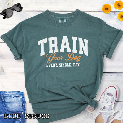 Green Comfort Colors t-shirt for dog training enthusiasts with 'TRAIN Your Dog EVERY. SINGLE. DAY.' text in bold athletic style lettering, shown on a wooden surface with jeans and shoes.