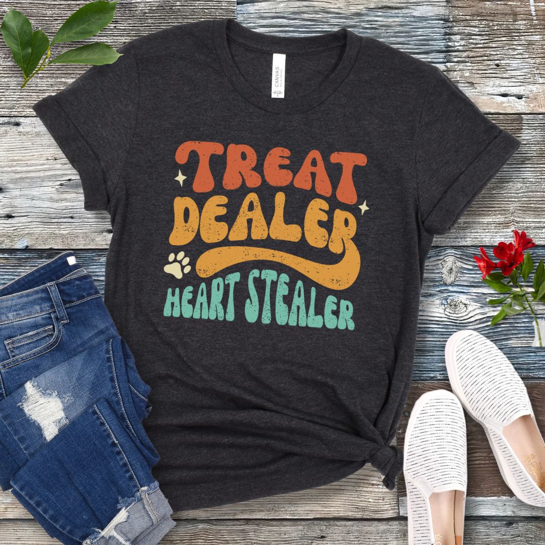 Dark grey t-shirt for dog lovers with the multicolor text 'TREAT DEALER HEART STEALER' printed in a groovy 70s-style font, accompanied by a paw print. The shirt is presented flat with a pair of blue jeans and white shoes.
