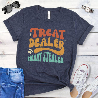 Heather navy t-shirt for dog lovers with the multicolor text 'TREAT DEALER HEART STEALER' printed in a groovy 70s-style font, accompanied by a paw print. The shirt is presented flat with a pair of blue jeans and white shoes.