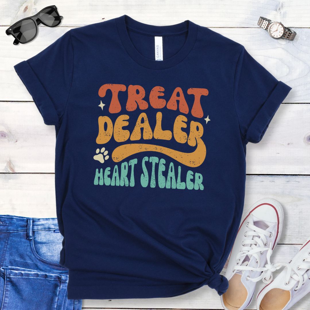 Navy t-shirt for dog lovers with the multicolor text 'TREAT DEALER HEART STEALER' printed in a groovy 70s-style font, accompanied by a paw print. The shirt is presented flat with a pair of blue jeans and white shoes.