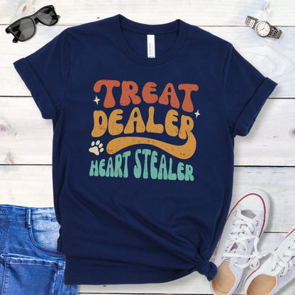 Navy t-shirt for dog lovers with the multicolor text 'TREAT DEALER HEART STEALER' printed in a groovy 70s-style font, accompanied by a paw print. The shirt is presented flat with a pair of blue jeans and white shoes.