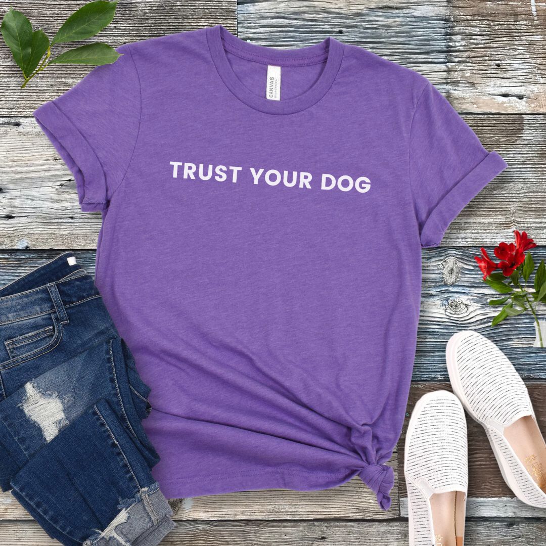 Purple dog training t-shirt with text 'Trust Your Dog' on a wooden surface with jeans and shoes.
