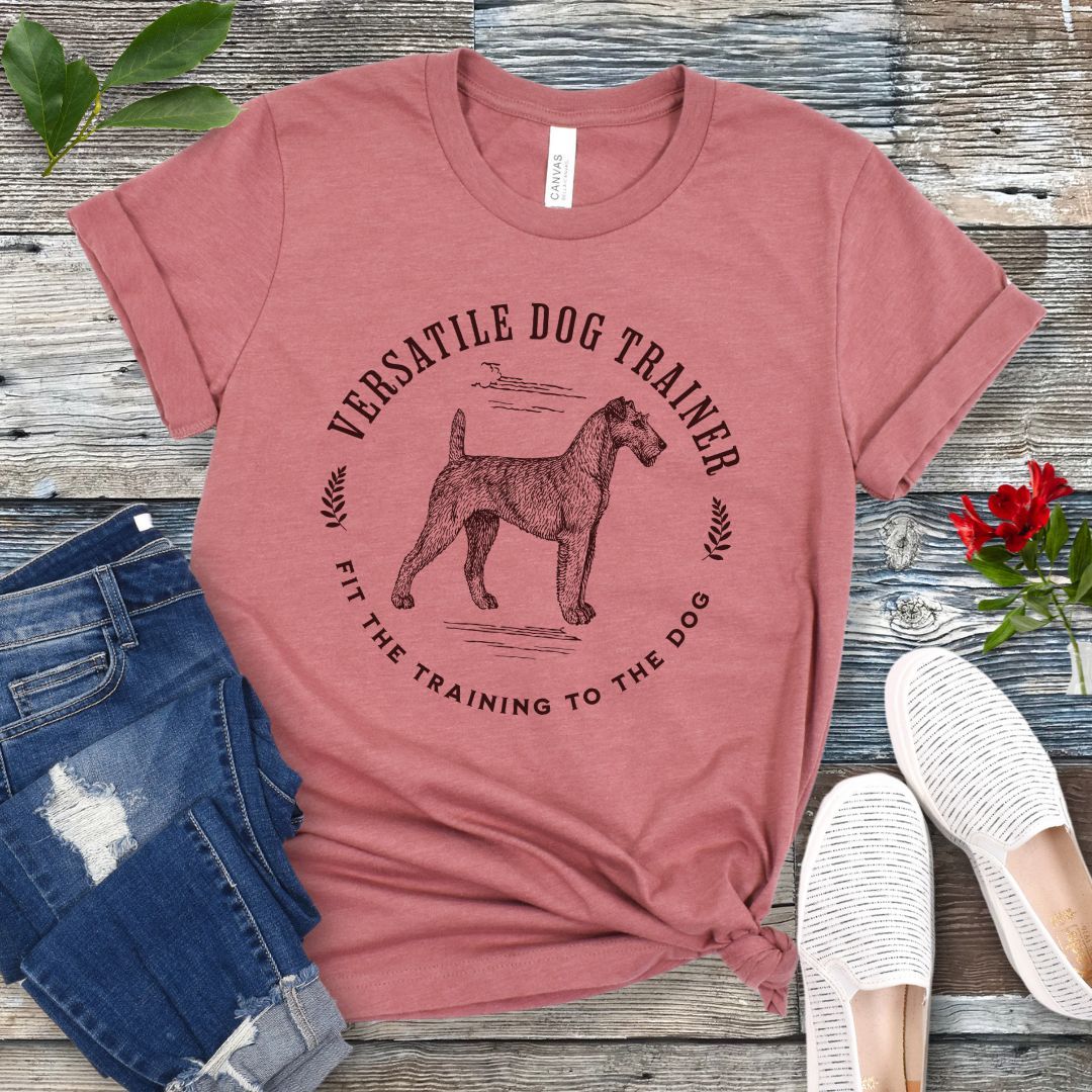 Mauve dog trainer it-shirt with vintage dog trainer graphic and text "Fit The Training To The Dog" on a wooden surface with jeans and shoes.