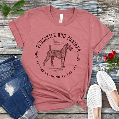 Mauve dog trainer it-shirt with vintage dog trainer graphic and text "Fit The Training To The Dog" on a wooden surface with jeans and shoes.