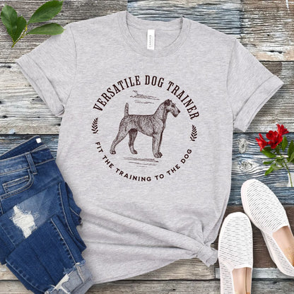 Light grey dog trainer it-shirt with vintage dog trainer graphic and text "Fit The Training To The Dog" on a wooden surface with jeans and shoes.