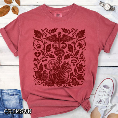 Crimson Comfort Colors Veterinary Medicine t-shirt with a monochrome linocut style design of a dog, a cat, and various veterinary and botanical elements, shown on a wooden surface with jeans and shoes.