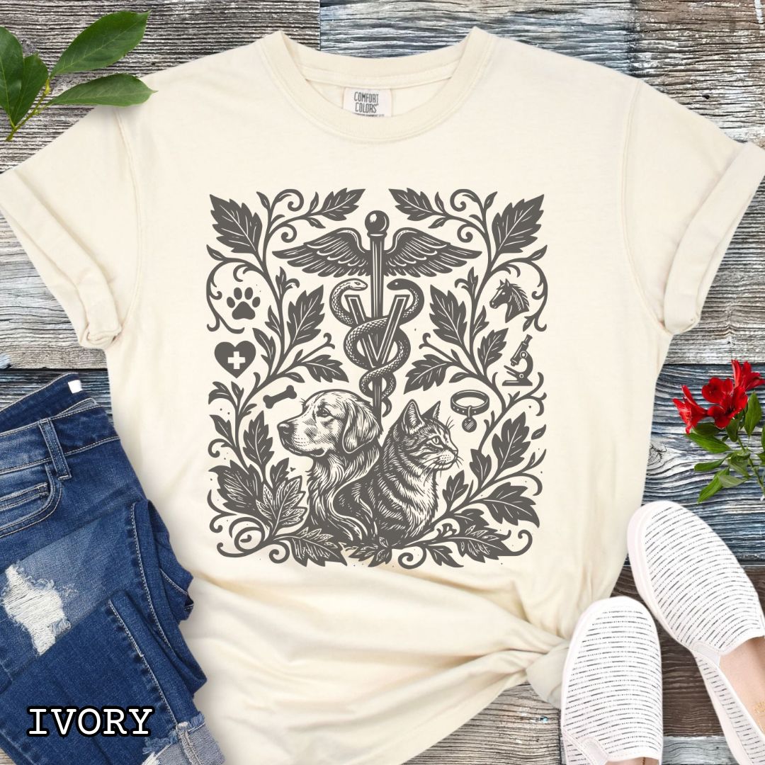 Beige Comfort Colors Veterinary Medicine t-shirt with a monochrome linocut style design of a dog, a cat, and various veterinary and botanical elements, shown on a wooden surface with jeans and shoes.