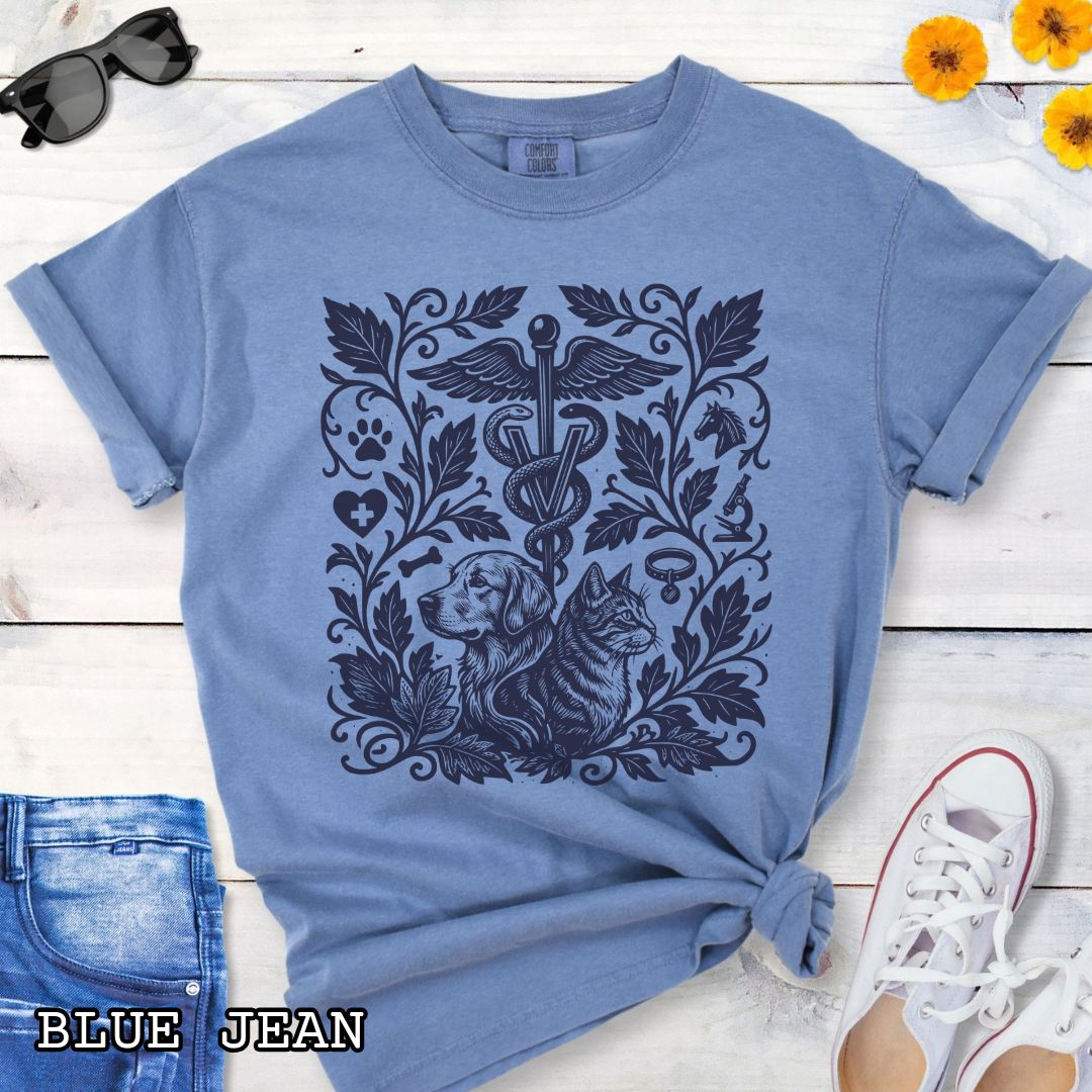 Blue Comfort Colors Veterinary Medicine t-shirt with a monochrome linocut style design of a dog, a cat, and various veterinary and botanical elements, shown on a wooden surface with jeans and shoes.
