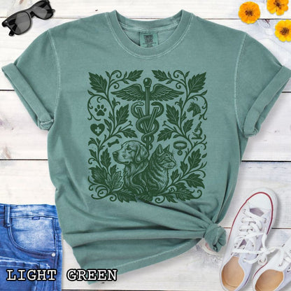 Green Comfort Colors Veterinary Medicine t-shirt with a monochrome linocut style design of a dog, a cat, and various veterinary and botanical elements, shown on a wooden surface with jeans and shoes.