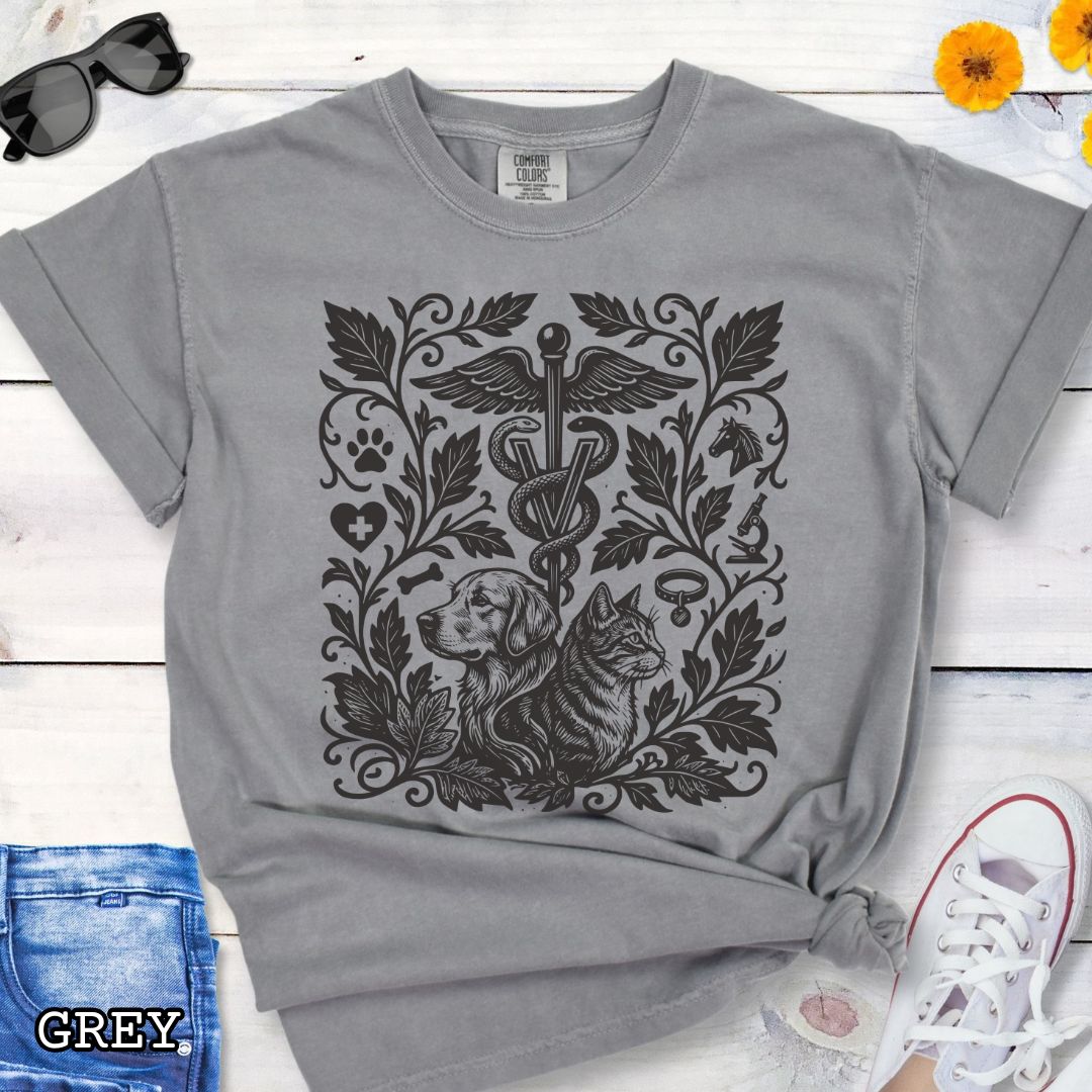 Grey Comfort Colors Veterinary Medicine t-shirt with a monochrome linocut style design of a dog, a cat, and various veterinary and botanical elements, shown on a wooden surface with jeans and shoes.