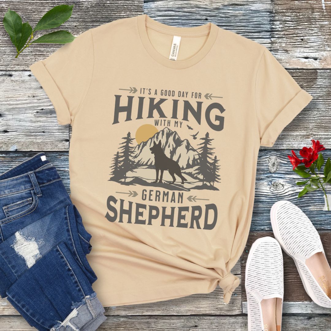 Cream t-shirt with vintage mountain scene, German Shepherd silhouette and 'Hiking with my Shepherd' text, presented on a wooden surface with jeans and shoes.