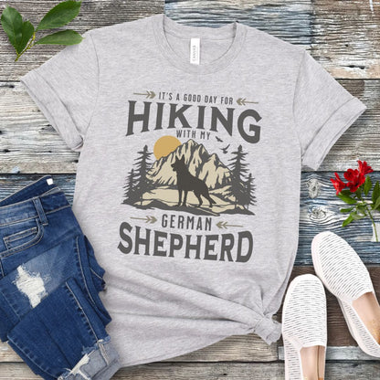 Light blue t-shirt with vintage mountain scene, German Shepherd silhouette and 'Hiking with my Shepherd' text, presented on a wooden surface with jeans and shoes.