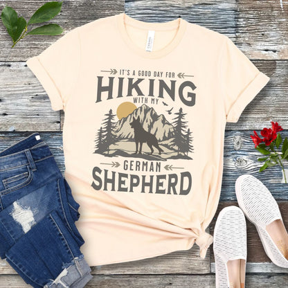 Beige t-shirt with vintage mountain scene, German Shepherd silhouette and 'Hiking with my Shepherd' text, presented on a wooden surface with jeans and shoes.