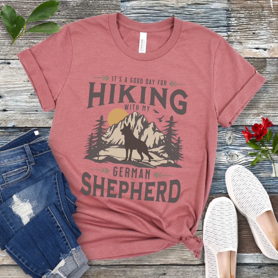 Mauve t-shirt with vintage mountain scene, German Shepherd silhouette and 'Hiking with my Shepherd' text, presented on a wooden surface with jeans and shoes.