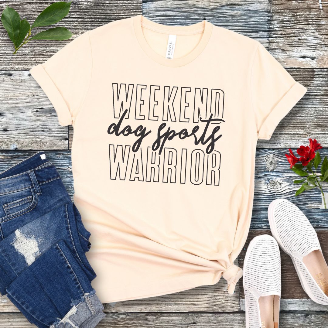 A beige dog training t-shirt with the text 'Weekend dog sports warrior' printed in black, displayed flat with a pair of blue jeans and white shoes.