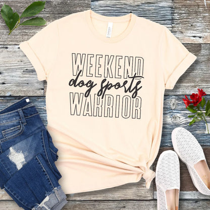 A beige dog training t-shirt with the text 'Weekend dog sports warrior' printed in black, displayed flat with a pair of blue jeans and white shoes.