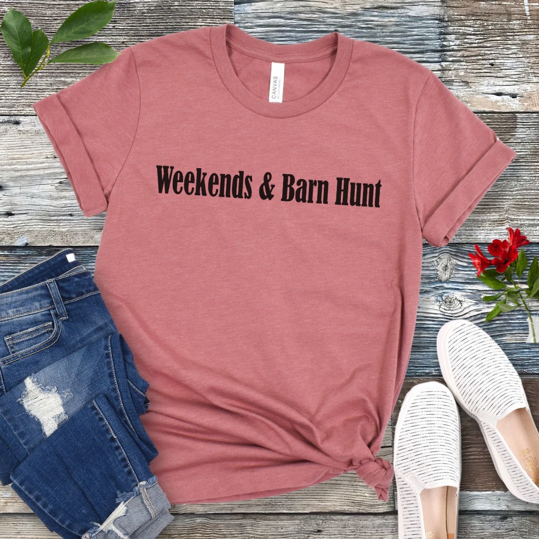 Mauve dog sports t-shirt with 'Weekends & Barn Hunt' text on a wooden surface with jeans and shoes.