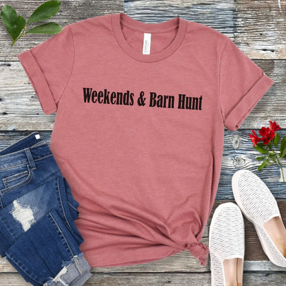 Mauve dog sports t-shirt with 'Weekends & Barn Hunt' text on a wooden surface with jeans and shoes.