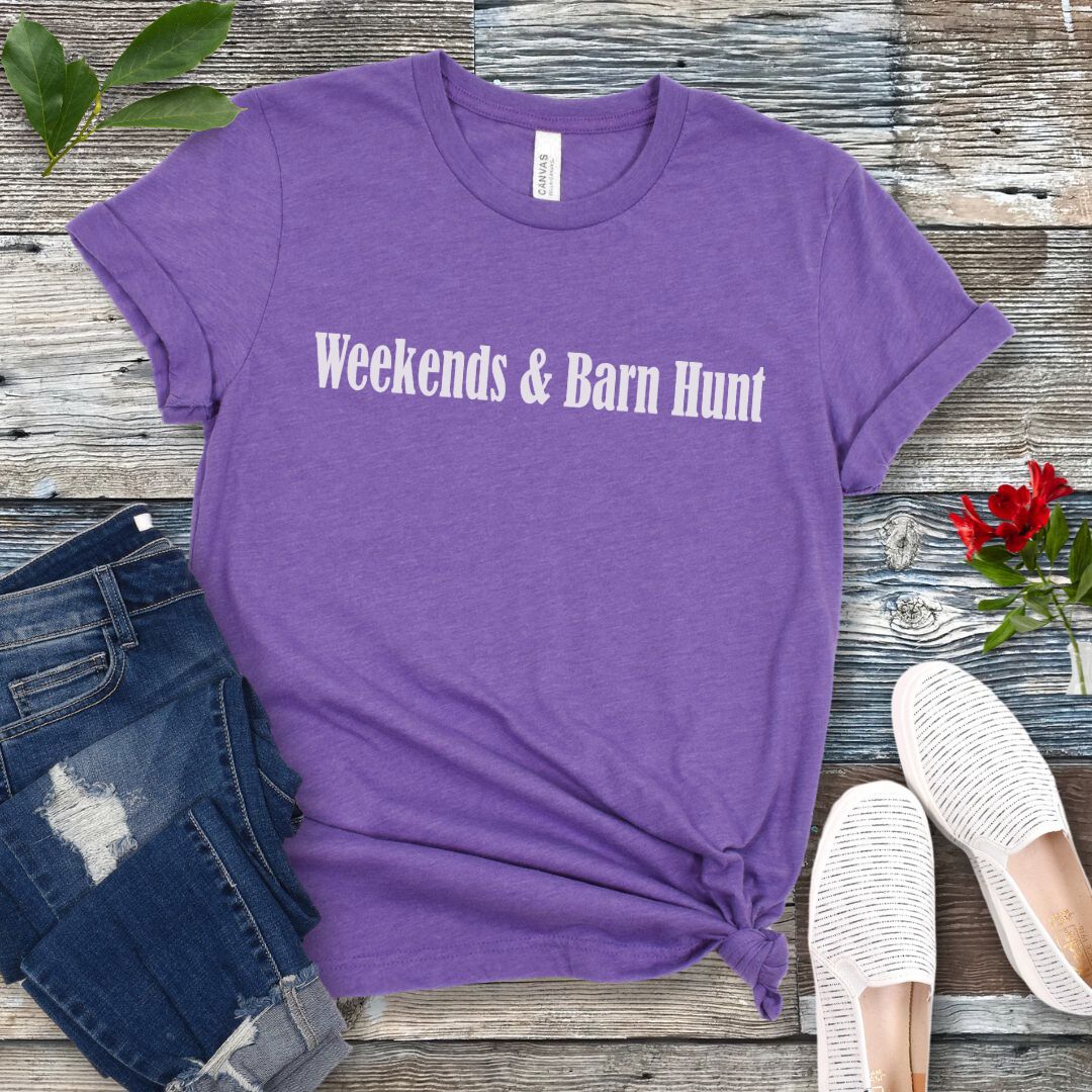 Purple dog sports t-shirt with 'Weekends & Barn Hunt' text on a wooden surface with jeans and shoes.