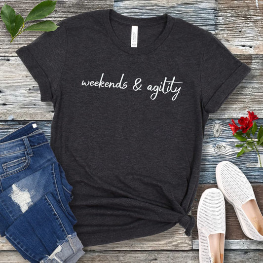 Dark grey dog agility t-shirt with 'weekends & agility' in script text on a wooden surface with jeans and shoes.